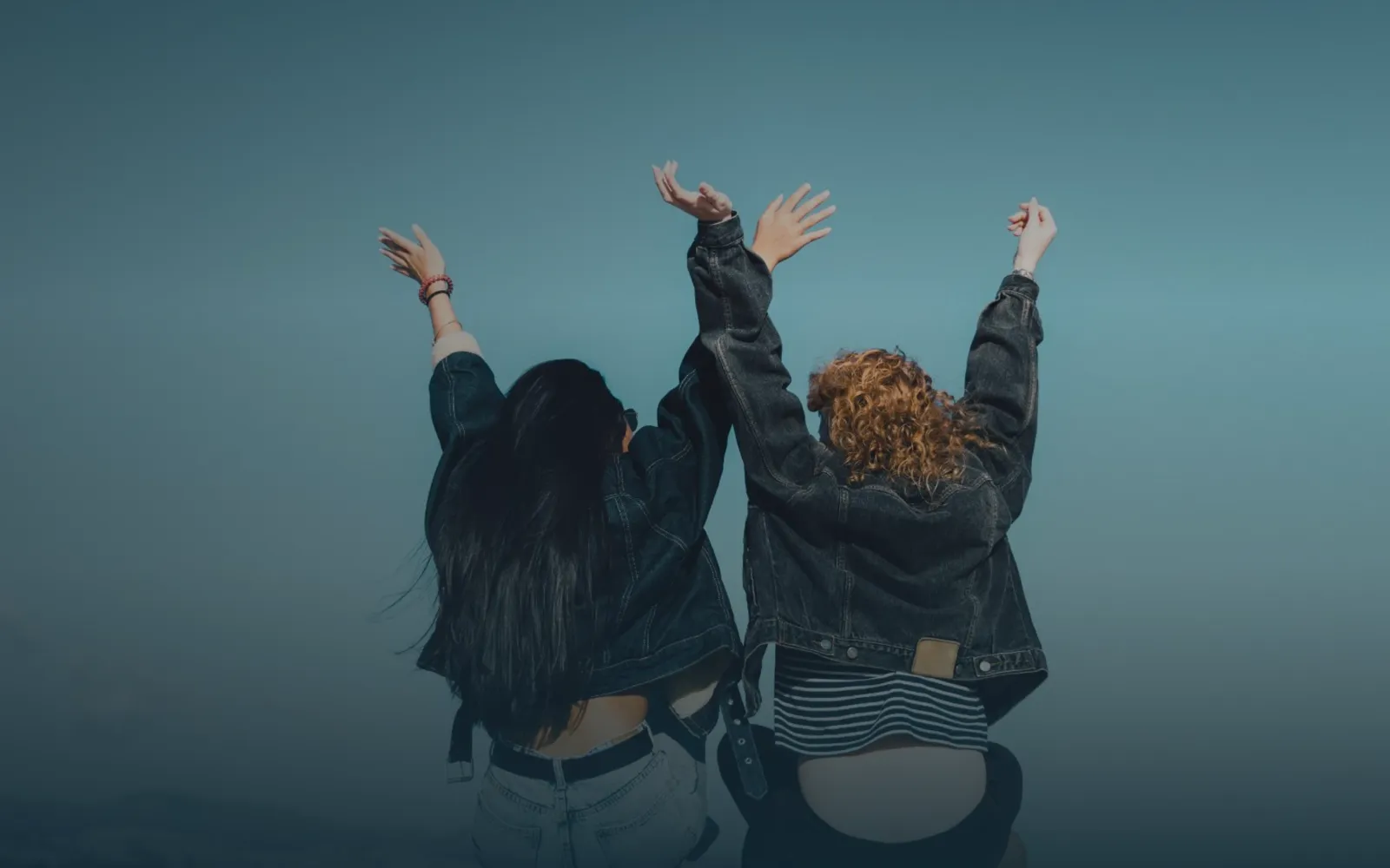 Dos jóvenes mujeres de espaldas, con chaquetas vaqueras, levantando los brazos hacia el cielo con entusiasmo, sobre un fondo azul grisáceo