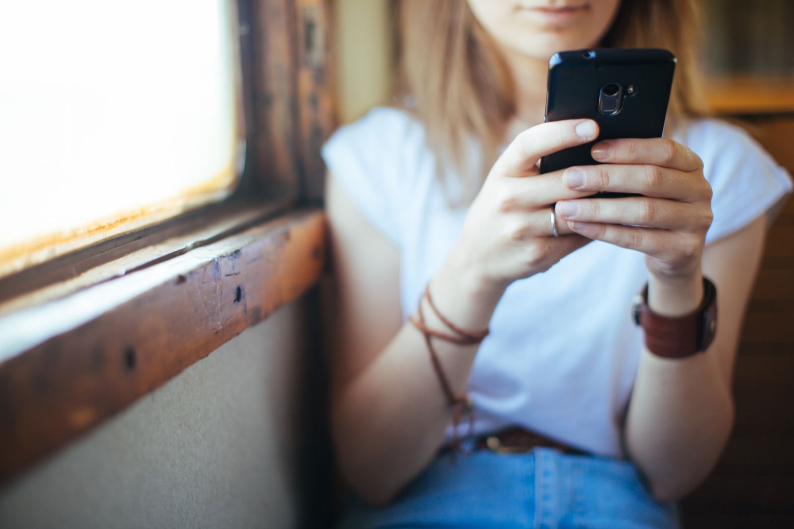 Young woman wearing a white T-shirt using her mobile phone while sitting next to a train window, with bracelets and a watch on her wrist.