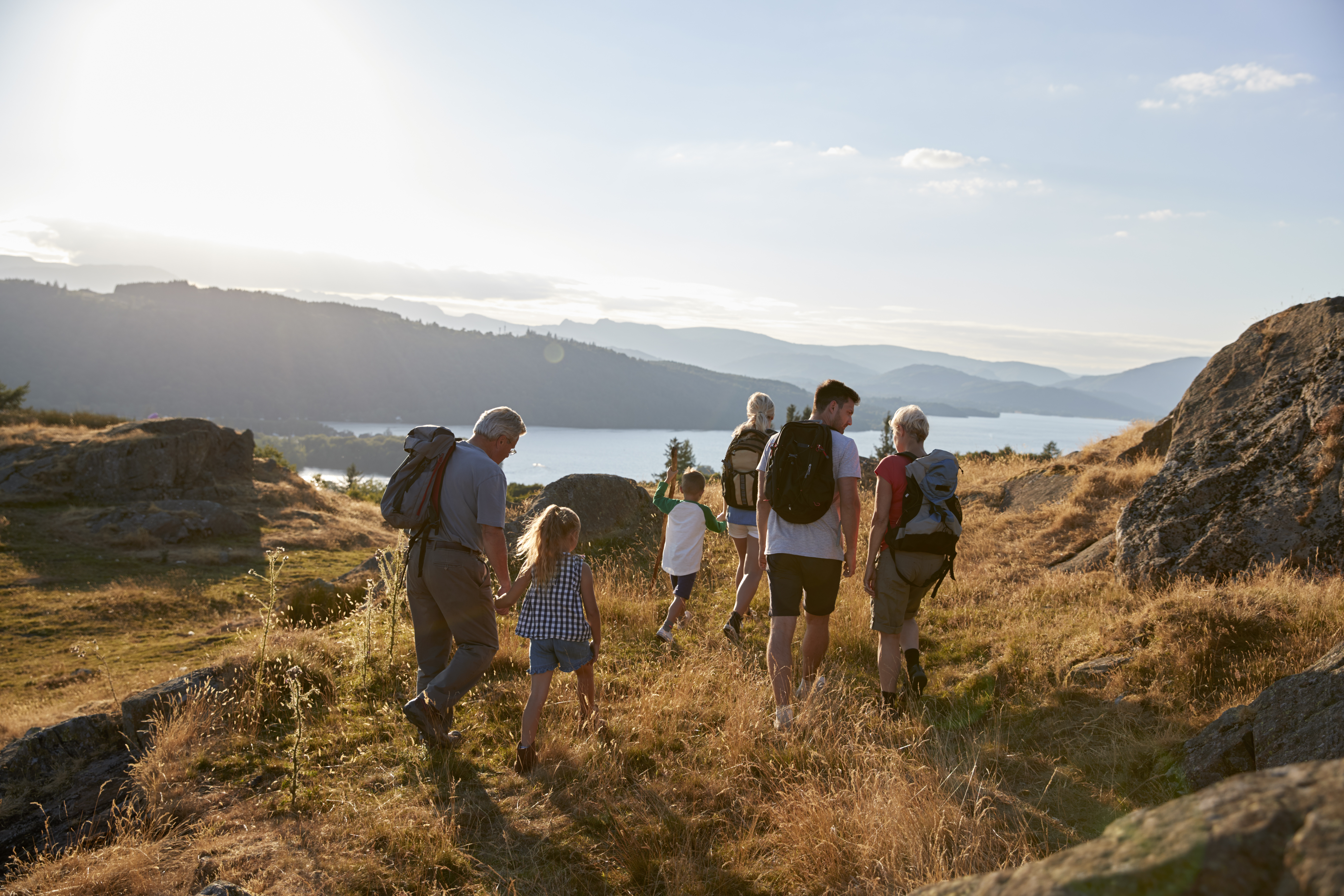Una familia multigeneracional de seis personas, incluyendo abuelos, padres e hijos con mochilas, camina de espaldas por un sendero de montaña dorado hacia un gran lago al atardecer. Imagen de fondo para un post sobre seguridad en viajes familiares de ellu.app